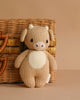 The Cuddle + Kind Baby Cow is a hand-knit beige plush made from heirloom-quality Peruvian cotton yarn, with white horns and a cream belly, pictured in front of a woven wicker basket on a light brown background.