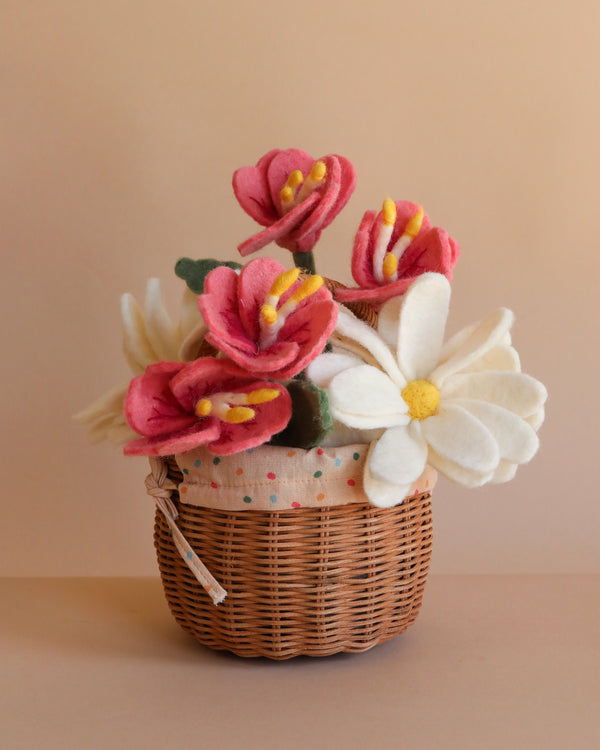 The Basket Of Felt Flowers features pink and white eco-friendly felt blooms with yellow centers, arranged in a small woven basket and displayed against a plain beige background.