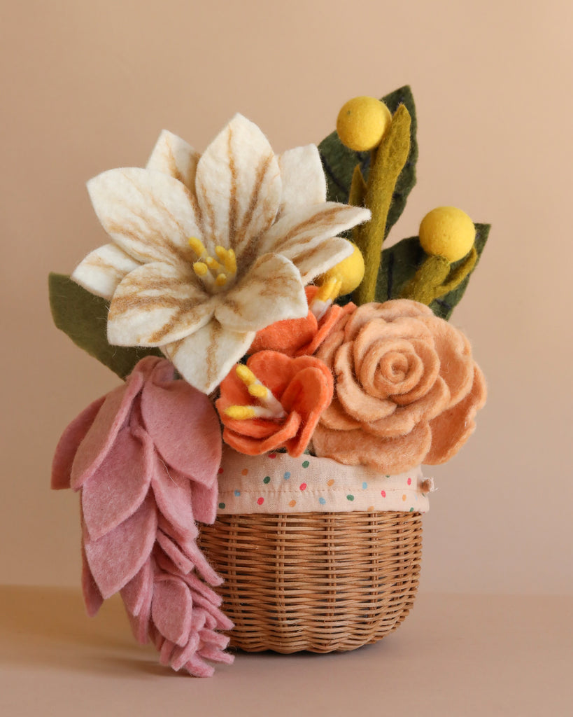 The Basket Of Felt Flowers features a woven basket with handcrafted felt blooms—large white and brown flower, peach roses, pink petals, yellow buds, and green leaves—displayed against a light beige background.