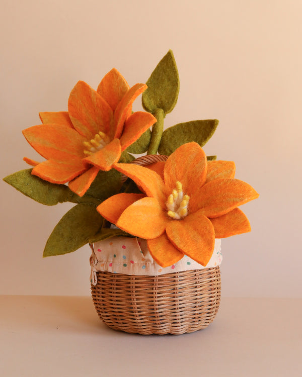 The Basket Of Felt Flowers features two large, bright orange handcrafted blooms with green felt leaves in a small wicker basket, displayed against a plain beige background.