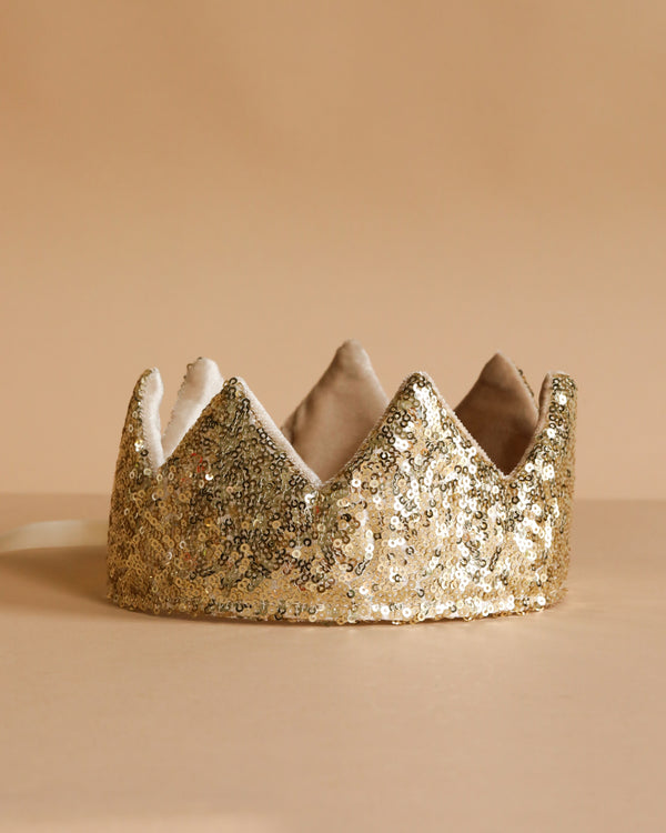 The Crown - Gold Sequins from Luciole et Petit Pois, inspired by France, features a pointed design with soft lining and sparkles brightly on a beige surface against a matching beige background.