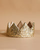 The Crown - Gold Sequins from Luciole et Petit Pois, inspired by France, features a pointed design with soft lining and sparkles brightly on a beige surface against a matching beige background.