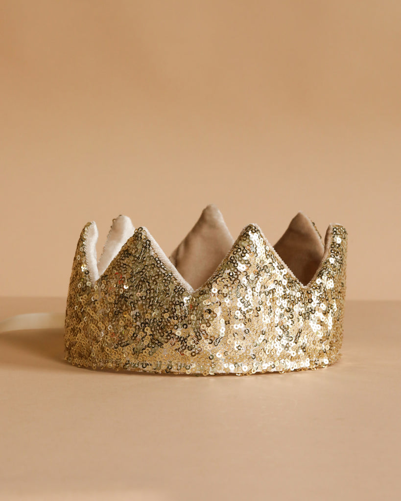 The Crown - Gold Sequins from Luciole et Petit Pois, inspired by France, features a pointed design with soft lining and sparkles brightly on a beige surface against a matching beige background.