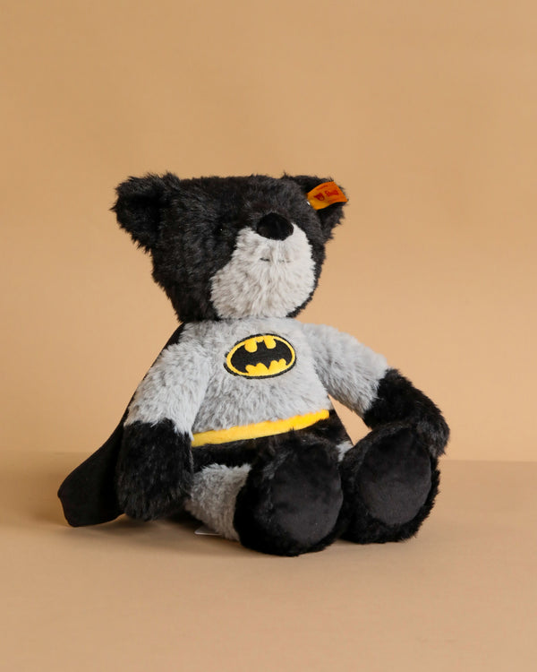 The 12-inch Steiff Batman Teddy Bear, dressed in a gray and black Dark Knight costume with a yellow belt and Batman logo, sits on a plain light brown background.