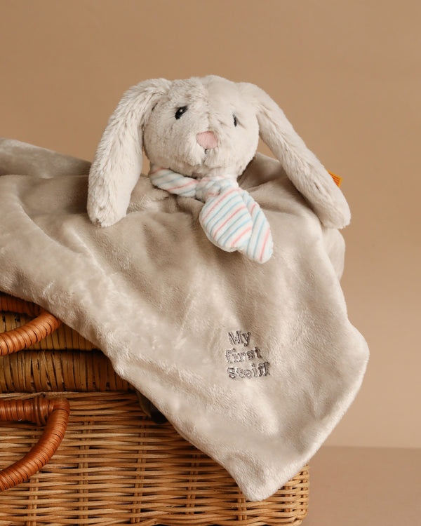 The Steiff Hoppie Rabbit Security Blanket Baby Toy, 10 Inches, features a soft beige blanket with a rabbit head, long ears, striped scarf, and "My first Steiff" embroidery—perfectly capturing classic Steiff charm. Draped elegantly over a wicker basket.