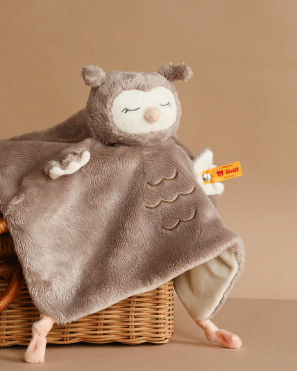 The Steiff Ollie Owl Baby Comforter, 10 inches, is a soft handcrafted plush toy with closed eyes, outstretched wings, and small ears. The signature Steiff button-in-ear tag is visible on its wing as it sits on a wicker basket against a beige background.