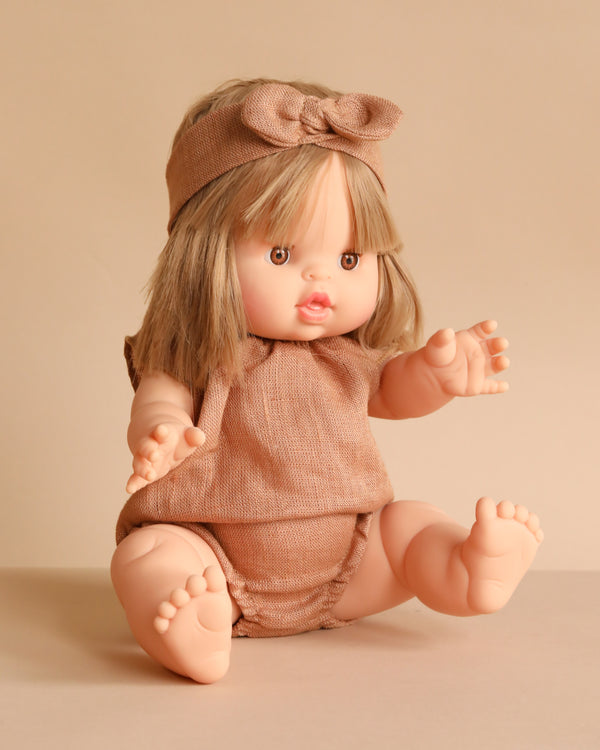 The Minikane Baby Doll (13") - Zoe With Clothing features light skin, straight blonde hair with bangs, a beige outfit, and a matching bow headband. This anatomically correct doll has outstretched arms and legs against a neutral beige background.