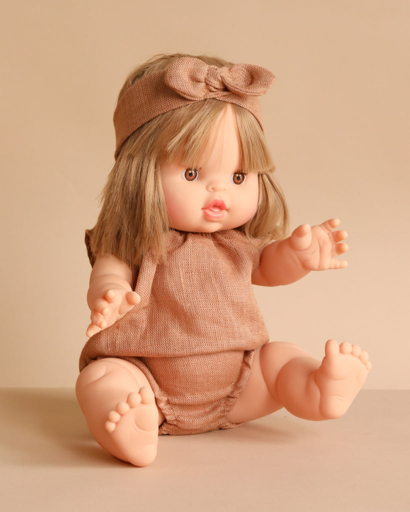 The Minikane Baby Doll (13") - Zoe With Clothing features light skin, straight blonde hair with bangs, a beige outfit, and a matching bow headband. This anatomically correct doll has outstretched arms and legs against a neutral beige background.