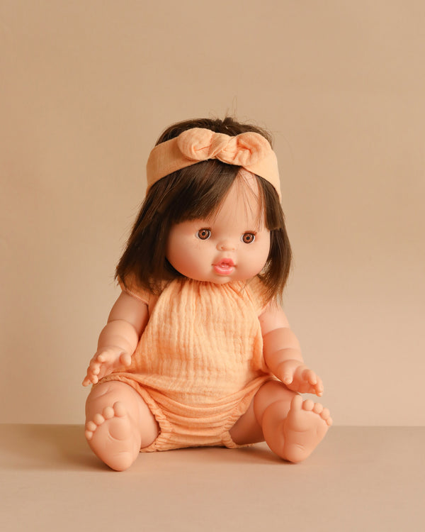 The Minikane Baby Doll (13") - Jeanne With Clothing features short brown hair, a peach headband with a bow, and a matching romper. This anatomically correct doll is ideal for imaginative play and is shown sitting on a beige surface against a beige background.