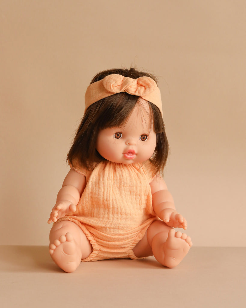 The Minikane Baby Doll (13") - Jeanne With Clothing features short brown hair, a peach headband with a bow, and a matching romper. This anatomically correct doll is ideal for imaginative play and is shown sitting on a beige surface against a beige background.