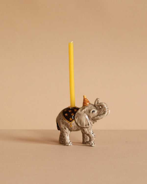 The Elephant Cake Topper features a porcelain elephant with a yellow candle on its back and an orange party hat, set against a beige background—an adorable collectible keepsake for any décor.