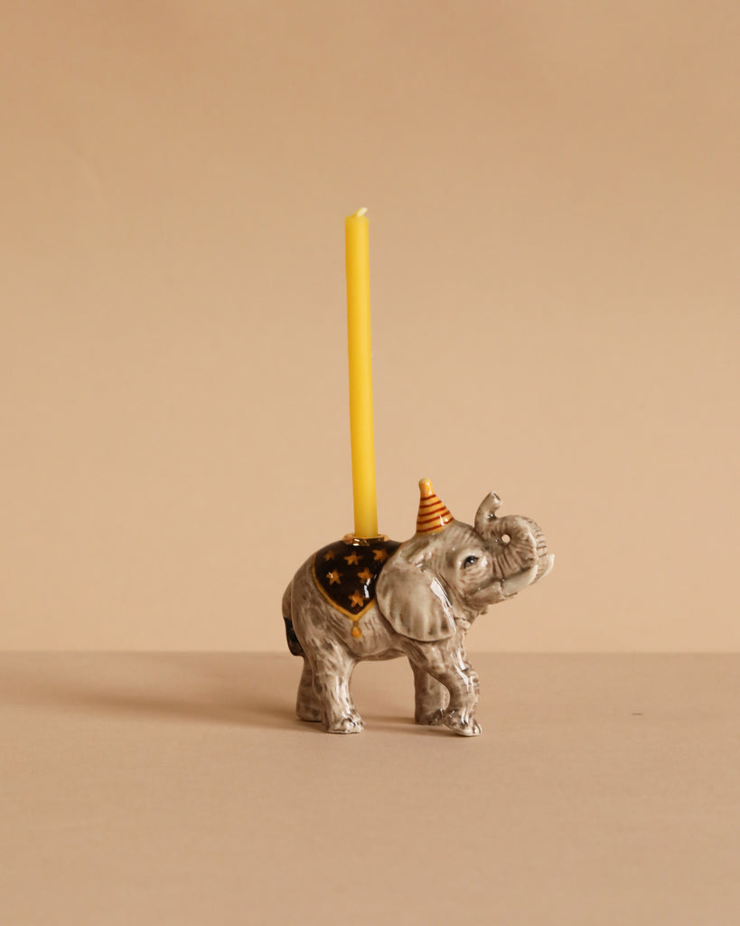 The Elephant Cake Topper features a porcelain elephant with a yellow candle on its back and an orange party hat, set against a beige background—an adorable collectible keepsake for any décor.