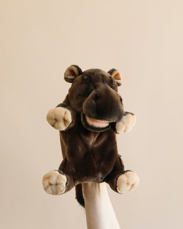 The Hippo Puppet, featuring a plush hand design, is showcased against a simple light grey backdrop. The puppet boasts a rich brown color with lighter beige accents on its feet, mouth, and nostrils. It also has plush ears, a wide mouth with a pink tongue, and small brown eyes that contribute to its charmingly realistic appearance.