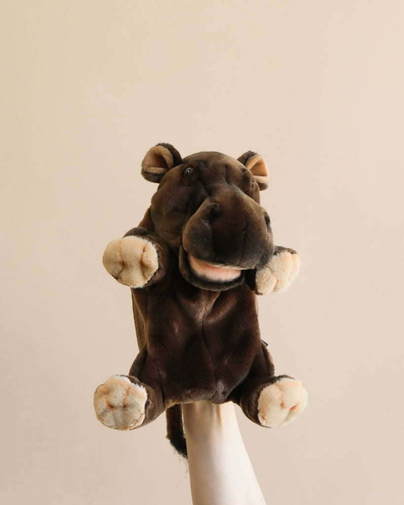 The Hippo Puppet, featuring a plush hand design, is showcased against a simple light grey backdrop. The puppet boasts a rich brown color with lighter beige accents on its feet, mouth, and nostrils. It also has plush ears, a wide mouth with a pink tongue, and small brown eyes that contribute to its charmingly realistic appearance.