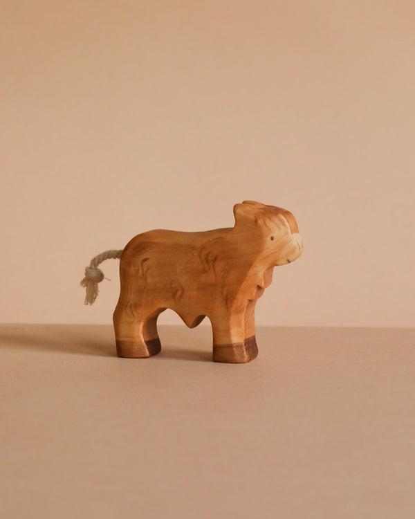 A small, handcrafted wooden toy named Handmade Wooden Highland Calf, made from sustainable hardwood with a minimalist design and short rope tail, is shown on a light brown surface against a beige background.
