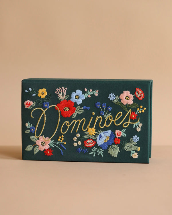 A dark green Strawberry Fields Dominoes Set keepsake box embroidered with "Dominoes" in gold script, featuring colorful illustrated flowers on a plain beige background.