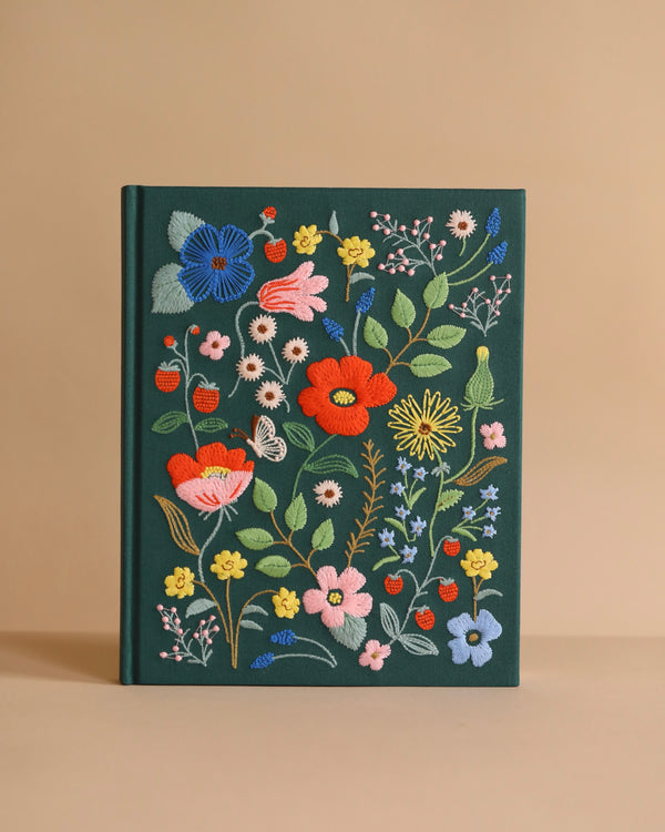 The Strawberry Fields Embroidered Sketchbook is a dark green hardcover journal with colorful floral, leaf, strawberry, and berry embroidery on the cover, displayed against a plain beige background.