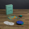 Board Game: Catch Phrase Vintage Bookshelf Edition displayed on a wooden surface, includes a teal box, scorecard, colorful markers, circular word disks, and a blue and black electronic game unit.