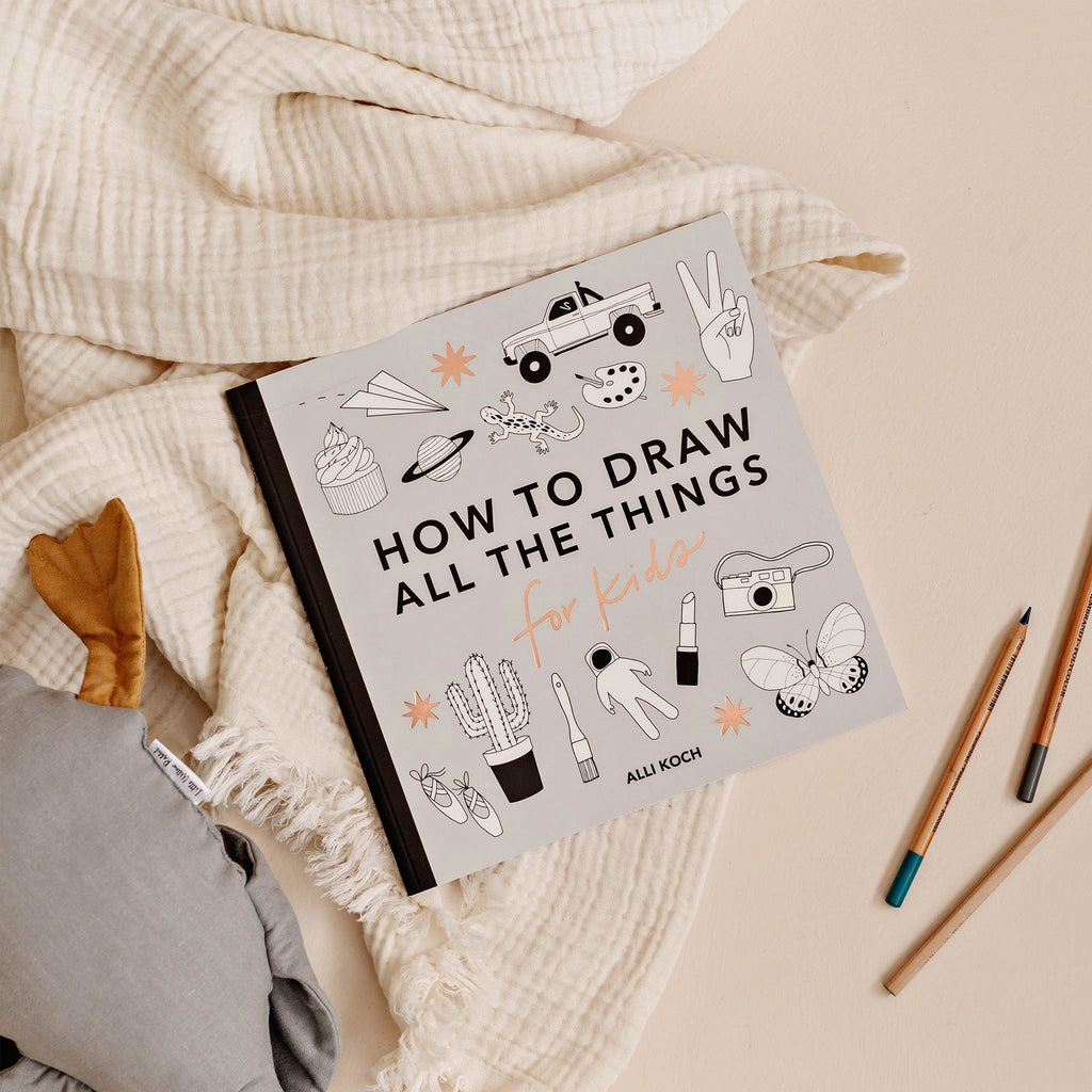 The “All the Things: How To Draw Books For Kids” art book sits on a cream blanket with colored pencils and an animal-footed plush toy. Its cover has simple line drawings, making it the perfect easy drawing book for children.