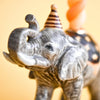 Close-up of the Elephant Cake Topper—a gray fine porcelain elephant figurine wearing a striped party hat, set against a blurred warm yellow background. An adorable collectible keepsake for your celebrations.
