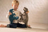 A young child sits on the floor, smiling and holding a Steiff Thaisen Tyrannosaurus "T-Rex" Dinosaur Plush Toy (14 inches) and a blue dino plush, with a white brick wall in the background.