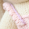 Close-up of Cuddle + Kind Briar The Bunny - Peony Pink shows hand knit cream and pink fabric with a textured cream background, a pink border, and thin lavender yarn, highlighting the detailed yarn and stitching.