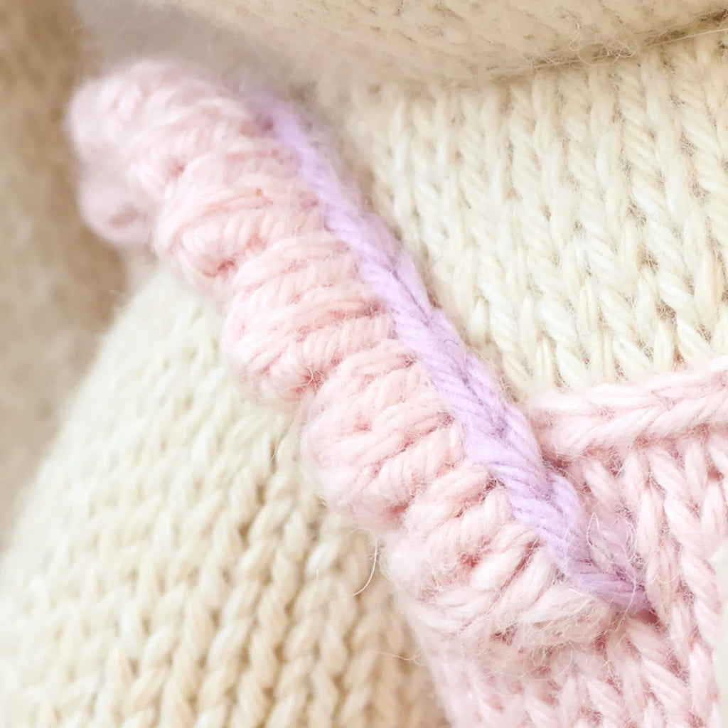 Close-up of Cuddle + Kind Briar The Bunny - Peony Pink shows hand knit cream and pink fabric with a textured cream background, a pink border, and thin lavender yarn, highlighting the detailed yarn and stitching.
