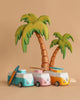 Three VW Beach Bus Sunset toy vans with surfboards are parked in front of two wooden palm trees, set against a plain beige background.