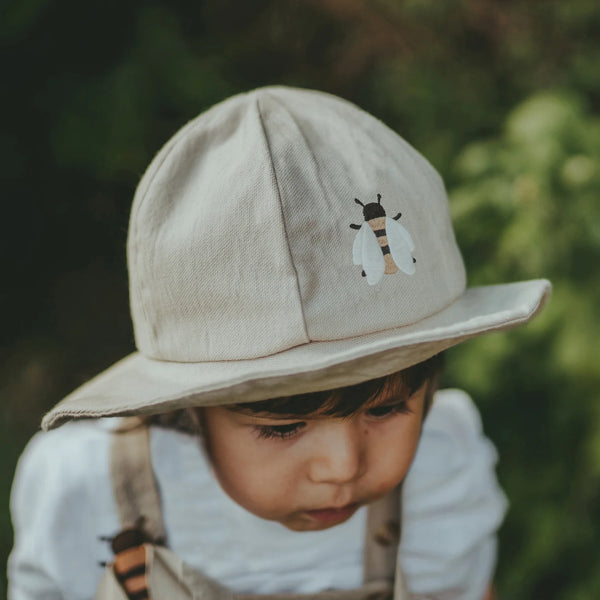 A young child looks downward, wearing the Donsje Organic Cotton Steijn Bee Hat. The light-colored hat features a bee graphic on the front and provides sun protection, set against blurred greenery in the background.