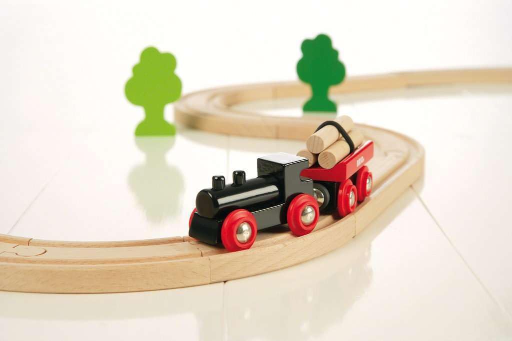 The BRIO Little Forest Train Set features a black engine and red log car on a curved wooden track, with two green tree figures in the background, all displayed on a white surface.