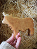 A hand holds the Handmade Wooden Highland Cow, a toy carved from sustainable hardwood, with straw or shredded wood in the background.
