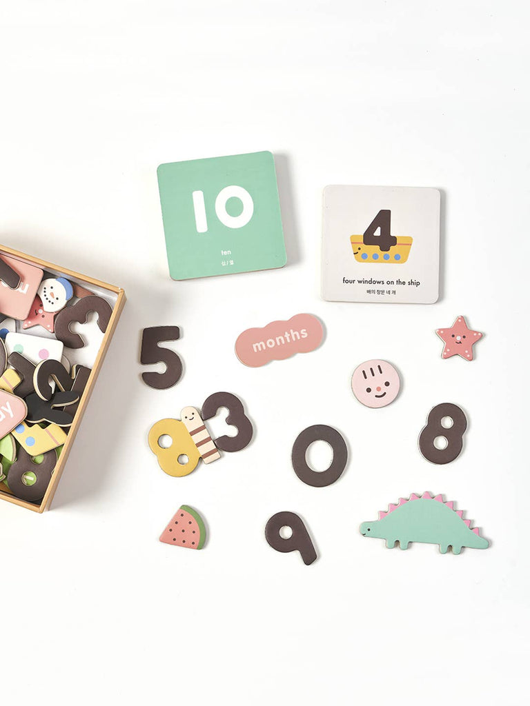 A flat lay of the Magnetic Number Play Set—colorful wooden numbers, animals, shapes, and two square cards ("10" and "4" with a boat)—arranged on a white surface beside an open box.