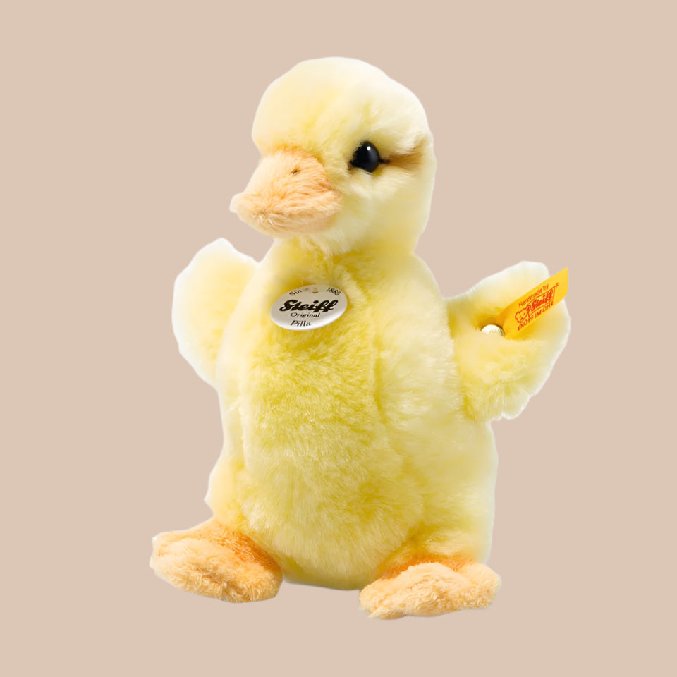 The Steiff Pilla Duckling Stuffed Animal is a plush yellow duckling with an orange beak and feet, Steiff chest tag, and yellow tag on its left wing—an ideal gift for collectors, shown on a plain beige background.