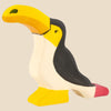 The Holztiger Toucan, handcrafted from maple and beech wood, features a yellow beak and foot, black back, white belly, and a pink tail detail—shown in classic HOLZTIGER style against a light beige background. Ships in one week.