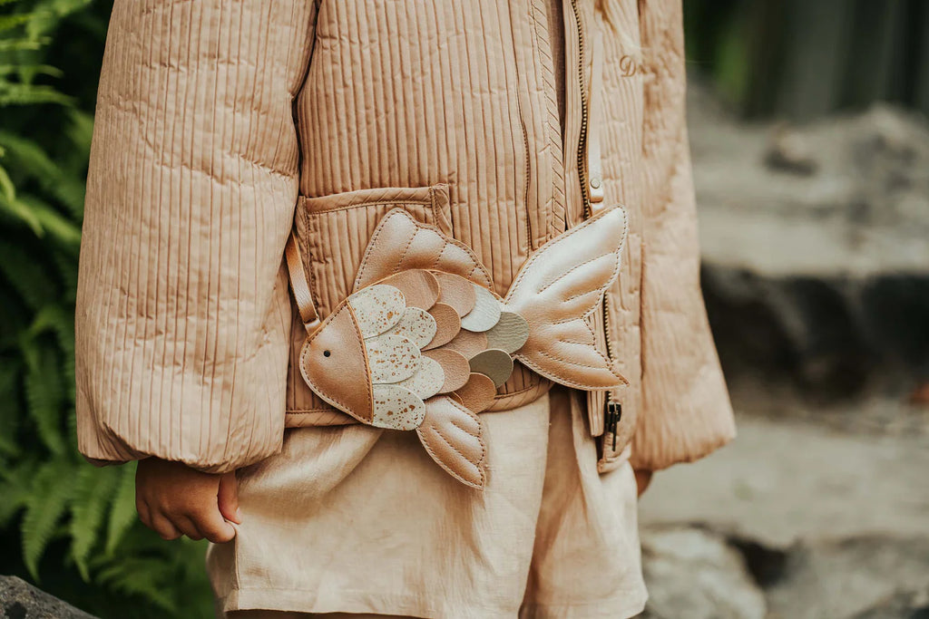 A child in a beige quilted jacket and shorts stands outside with the Donsje Leather Thysa Purse - Gold Fish, a fish-shaped bag featuring textured, multicolored scales and an adjustable strap. Ferns and rocks are in the background.