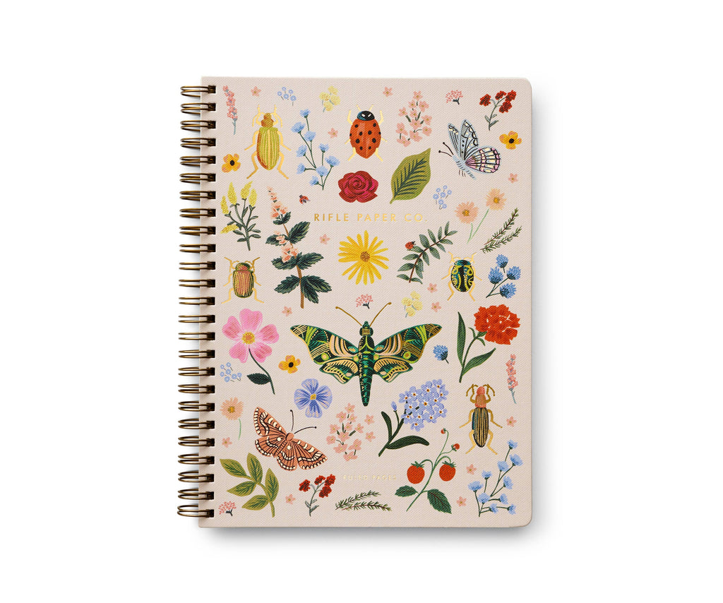 The Curio Spiral Notebook is a spiral-bound softcover with a cream cover featuring colorful florals, insects, and strawberries. "Rifle Paper Co." is printed in gold at the center. It includes ruled pages for easy writing.