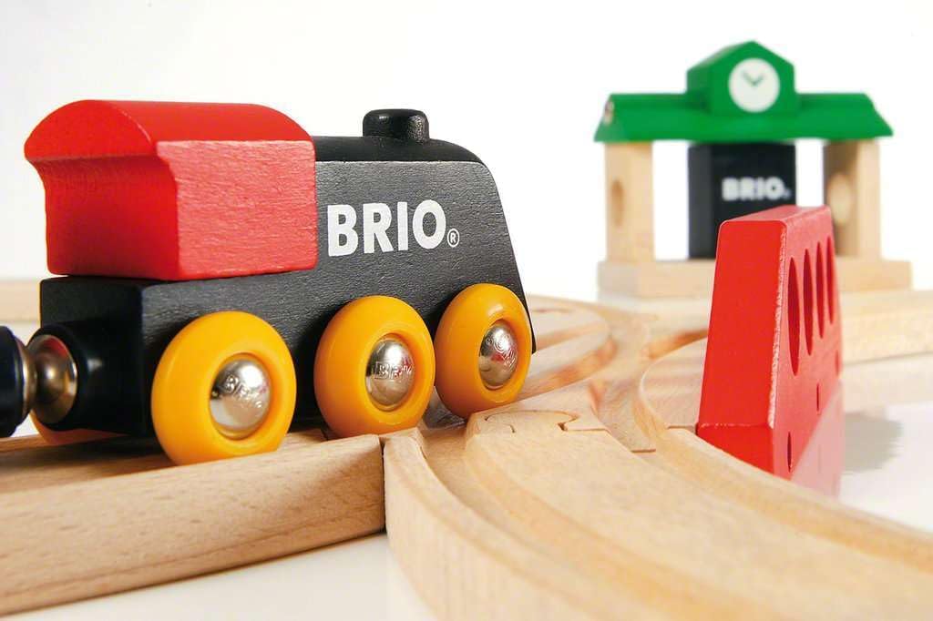A close-up of the BRIO World Train Set Classic Figure 8 Set shows a wooden toy train with red and black colors, yellow wheels, and a red bumper on its track. A small green and white station building appears in the background.