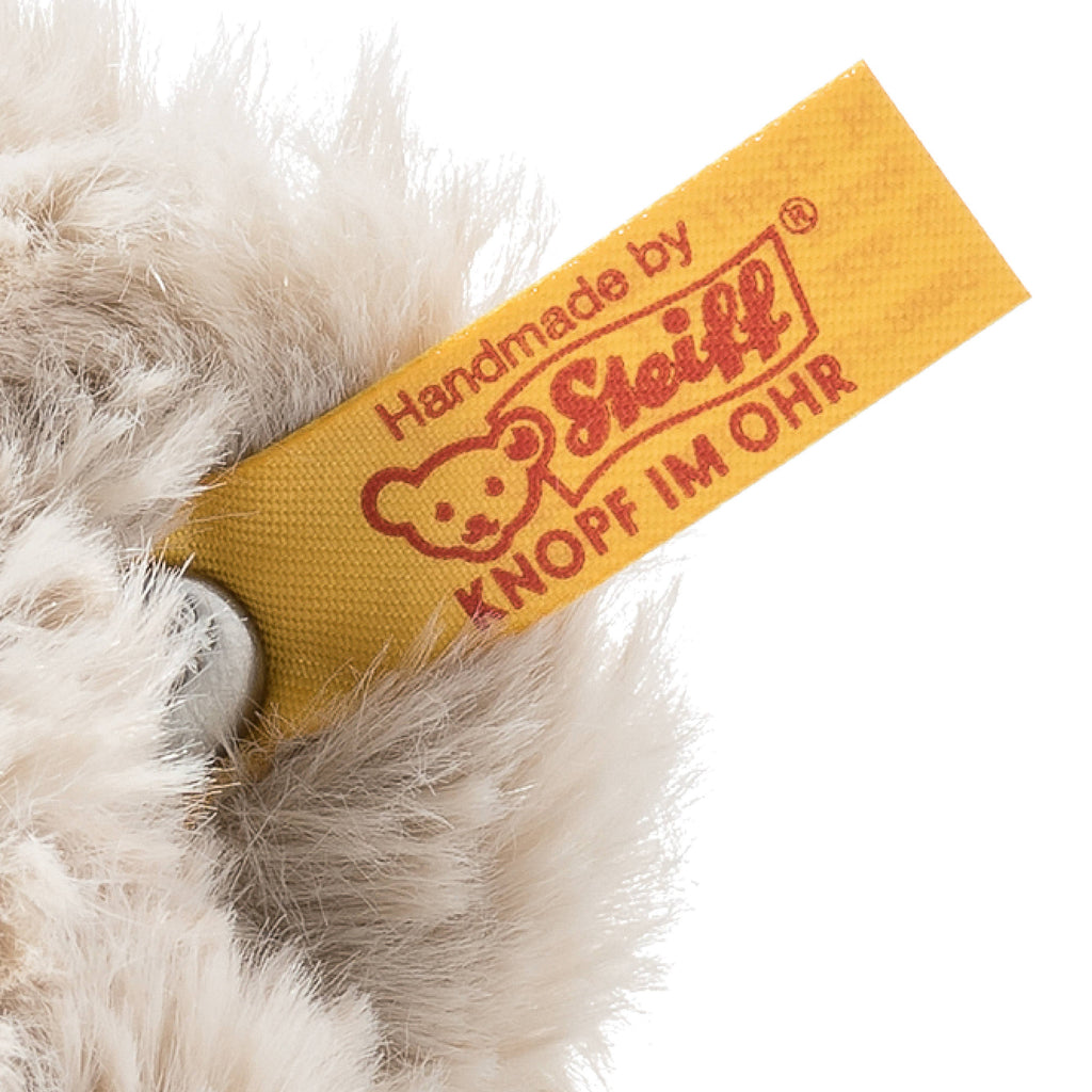 Close-up of the ear of the Steiff Honey Teddy Bear Stuffed Plush, 8 Inches, showing its yellow "Handmade by Steiff KNOPF IM OHR" tag with bear face logo and signature metal button—iconic features of this beloved toy.