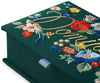 A close-up of the Strawberry Fields Dominoes Set, an embroidered keepsake box in green fabric with colorful flower designs, gold cursive writing, and a blue ribbon tab at the edge.