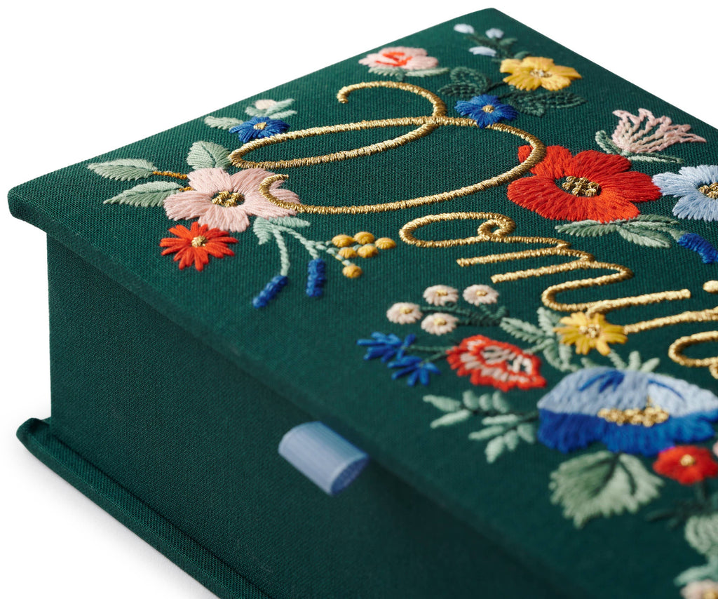 A close-up of the Strawberry Fields Dominoes Set, an embroidered keepsake box in green fabric with colorful flower designs, gold cursive writing, and a blue ribbon tab at the edge.