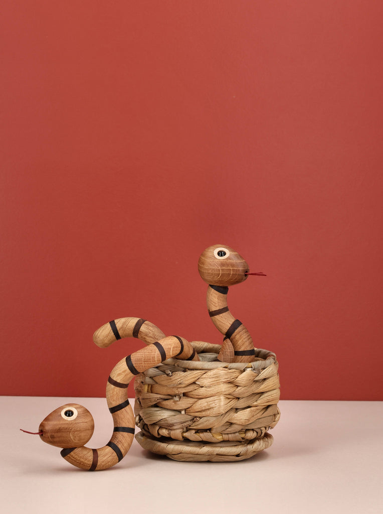 Two Spring Copenhagen The Snake wooden figurines with painted eyes and tongues are displayed in and beside a woven basket against a muted terracotta background—one coiled on the surface, the other rising from the basket—a unique spiritual gift.