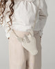 A child in a white blouse and beige pants holds the Donsje Leather Blinc Purse - Unicorn, a white leather purse with ears, horn, and an adjustable strap for comfort. Curly hair falls to one side.