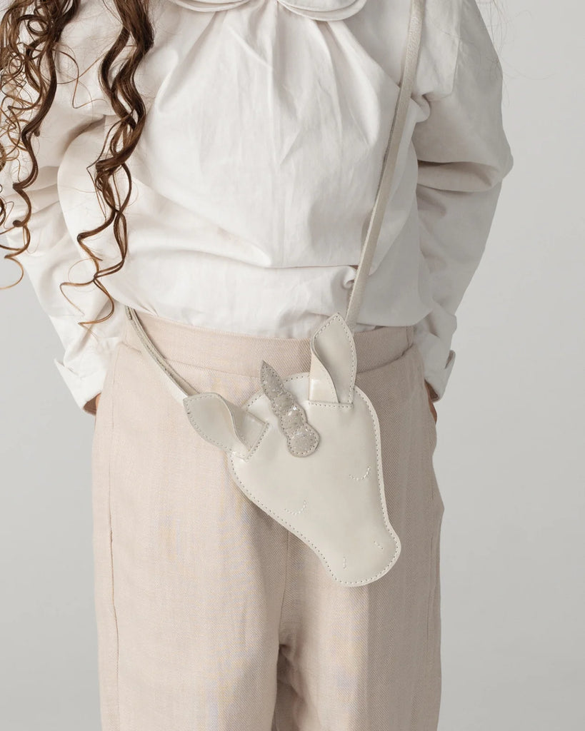A child in a white blouse and beige pants holds the Donsje Leather Blinc Purse - Unicorn, a white leather purse with ears, horn, and an adjustable strap for comfort. Curly hair falls to one side.
