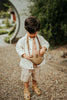 A young boy with brown hair stands on a pebble path, wearing a white jacket, light brown shirt and shorts, tan sandals, and carrying the Donsje Leather Talei Bumbag Purse - Sloth. Greenery and a round entryway are in the background.