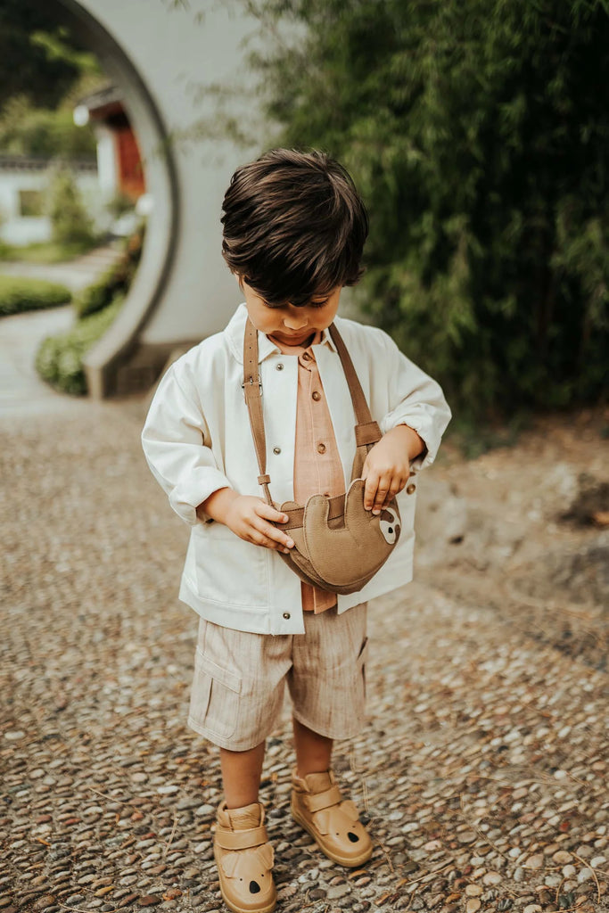 A young boy with brown hair stands on a pebble path, wearing a white jacket, light brown shirt and shorts, tan sandals, and carrying the Donsje Leather Talei Bumbag Purse - Sloth. Greenery and a round entryway are in the background.