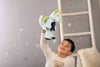 A young boy in a white bear-print shirt holds up the Steiff Samu The Magic Dragon Plush Toy, 11 Inches, in a cozy, star-decorated room with a white ladder and fairy lights—a legendary gift for any child.