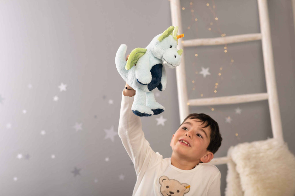 A young boy in a white bear-print shirt holds up the Steiff Samu The Magic Dragon Plush Toy, 11 Inches, in a cozy, star-decorated room with a white ladder and fairy lights—a legendary gift for any child.
