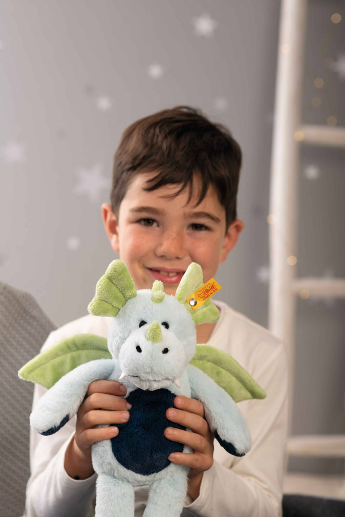 A smiling young boy with brown hair holds a Steiff Samu The Magic Dragon Plush Toy, 11 inches, towards the camera. A softly blurred background with white star decorations on a gray wall highlights this legendary gift.
