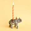 The Elephant Cake Topper is a fine porcelain figurine wearing a party hat and star-patterned blanket, holding a lit twisted orange candle on its back—an heirloom collectible displayed on a solid yellow background.