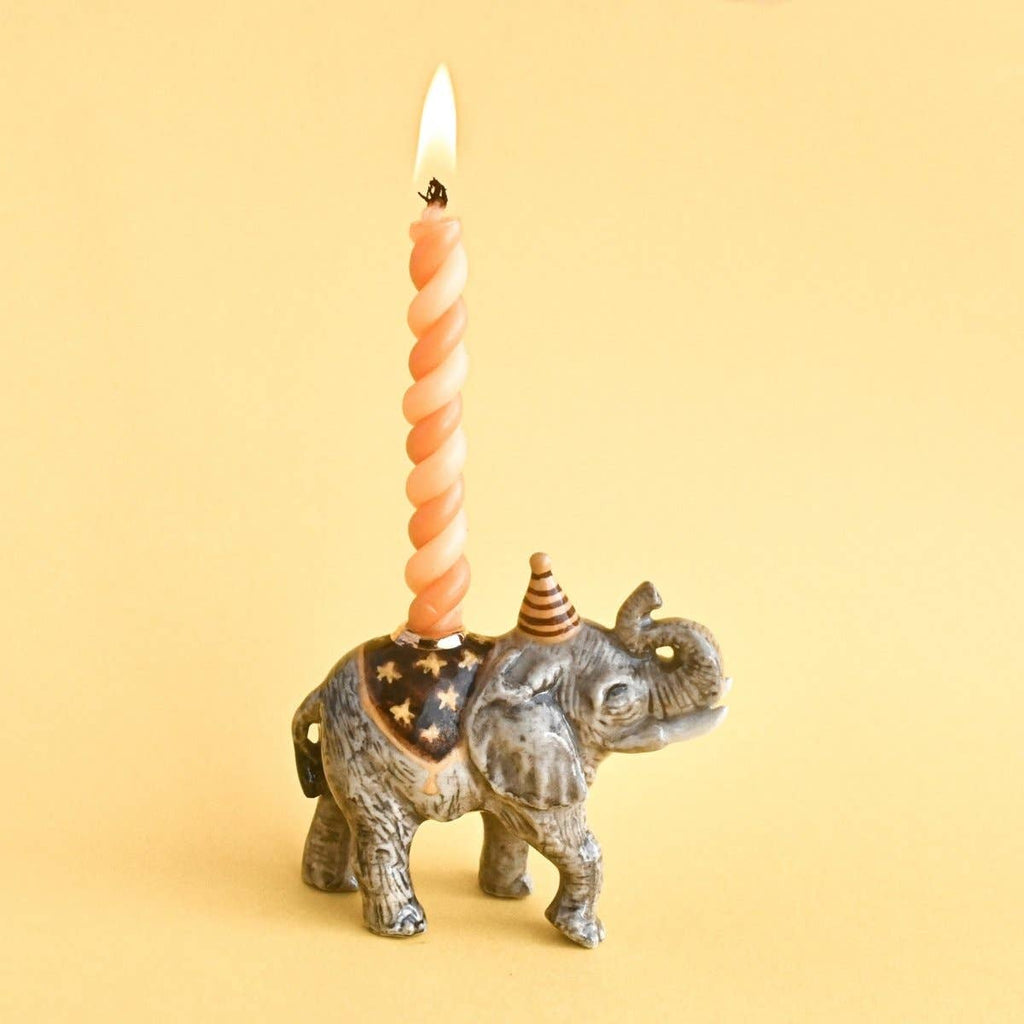The Elephant Cake Topper is a fine porcelain figurine wearing a party hat and star-patterned blanket, holding a lit twisted orange candle on its back—an heirloom collectible displayed on a solid yellow background.